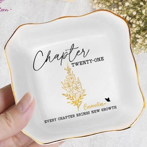 May include: White ceramic trinket dish with gold trim. The dish features the words "Chapter Twenty-One" and "Every Chapter Brings New Growth" in black script. A gold floral design and the name "Camelia" are also present.