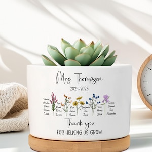 May include: A white ceramic planter with a succulent plant, personalized with the text "Mrs. Thompson 2024-2025" and student names. The planter sits on a wooden base, with the words "Thank you for helping us grow". A wooden clock is in the background.