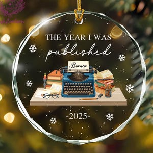 May include: A clear, round Christmas ornament with the text "THE YEAR I WAS published -2025". The ornament features a blue typewriter, books, and snowflakes. A name "Bianca" is on the typewriter.