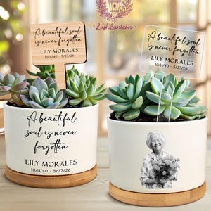 May include: Two white ceramic planters on bamboo bases, each holding a succulent. One features a wooden stake and text reading A beautiful soul is never forgotten, LILY MORALES, 10/11/40 - 5/27/26. The other has a clear acrylic stake and a photo.