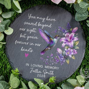 May include: A round, dark gray slate memorial stone with a hummingbird and floral design. The text reads "You may have soared beyond our sight, but you're forever in our hearts." Includes "In Loving Memory" and a name and year.