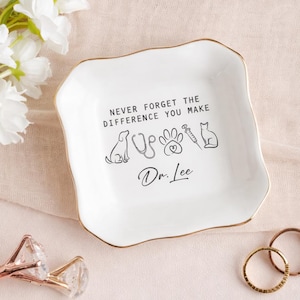 May include: White ceramic trinket dish with a gold rim. The dish features the text "NEVER FORGET THE DIFFERENCE YOU MAKE" and illustrations of a dog, stethoscope, paw print, syringe, and cat. The name "Dr. Lee" is also printed on the dish.