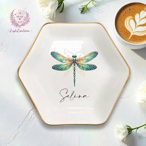 May include: A white hexagonal ceramic trinket dish with a gold-toned rim. The centre features a colourful dragonfly illustration with iridescent blue, green, and orange wings. The name Selina is printed in black cursive script below the dragonfly.
