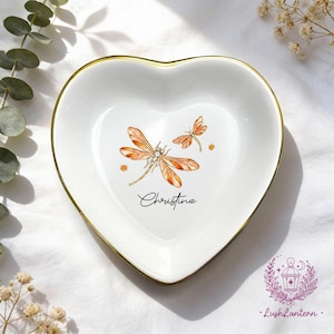 May include: White heart-shaped trinket dish with a gold rim, featuring two orange dragonflies and the name "Christine" in script. The dish is decorated with small star-like accents.