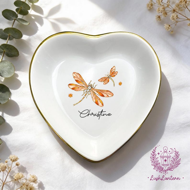 May include: White heart-shaped trinket dish with a gold rim, featuring two orange dragonflies and the name "Christine" in script. The dish is decorated with small star-like accents.