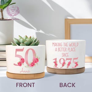 May include: Two white ceramic planters with bamboo saucers. One planter displays the number "50" in pink with floral accents and the name "Anna". The other reads "MAKING THE WORLD A BETTER PLACE SINCE 1975".