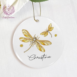 May include: White ceramic ornament featuring two yellow and gold dragonflies with crystal accents. The ornament has a gold hanging cord and the name "Christine" in script. Decorative floral accents are also present.