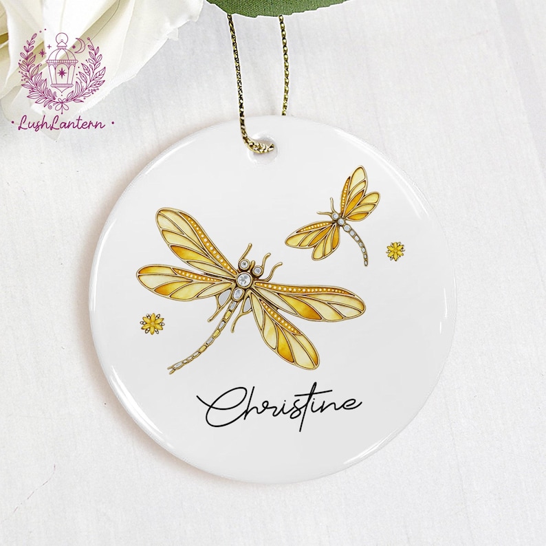 May include: White ceramic ornament featuring two yellow and gold dragonflies with crystal accents. The ornament has a gold hanging cord and the name "Christine" in script. Decorative floral accents are also present.