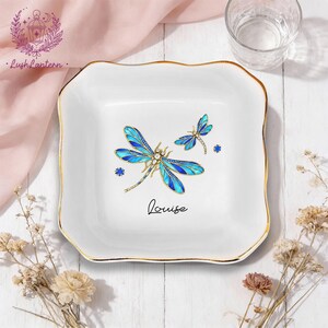 May include: White square trinket dish with a gold rim and a blue dragonfly design. The dish has the name "Louise" written in cursive. The dragonfly has blue wings and gold accents.