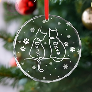 May include: Clear, faceted glass Christmas ornament with a red ribbon. The ornament features two cat silhouettes, the names "Doris" and "Dave", paw prints, and snowflakes. A festive decoration for cat lovers.