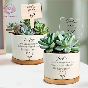 May include: Two succulent plants in white ceramic pots with wooden bases. Each pot has a name and a message printed on it, along with a stethoscope heart design. One plant has a wooden sign, the other a clear sign.