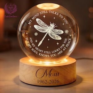 May include: A crystal ball night light with a dragonfly design and the text "MY MIND STILL TALKS TO YOU" and "Mom 1962-2026". The light sits on a wooden base, creating a warm ambiance. The base is approximately 7 cm in diameter.