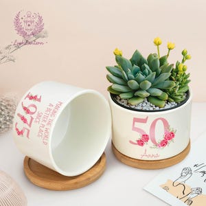 May include: Two white ceramic planters with succulents. One planter has the number "50" with floral accents and the name "Anna". The other planter has the text "1975 Since Making the World a Better Place". Both planters sit on wooden coasters.