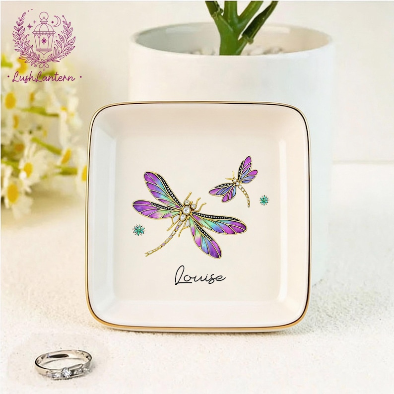 May include: Square, off-white trinket dish with a gold rim, featuring two dragonfly illustrations in purple, green, and gold. The name "Louise" is written in cursive. A silver ring with three stones sits in front of the dish.