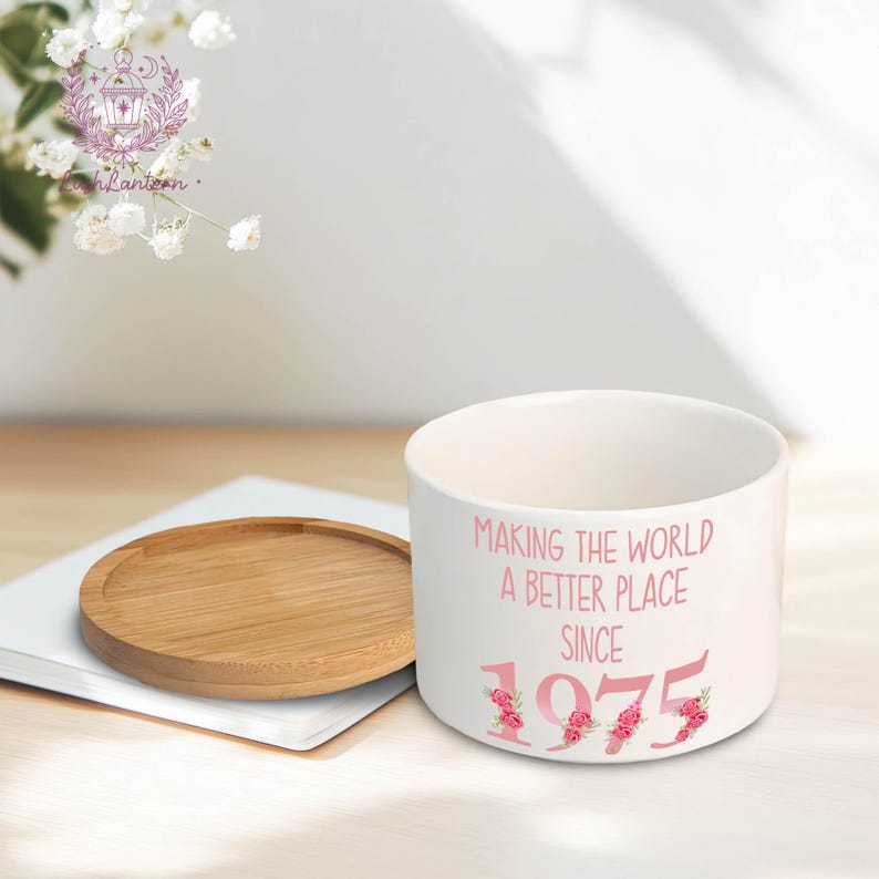 May include: A white ceramic planter with the text "MAKING THE WORLD A BETTER PLACE SINCE 1975" in pink. A round, wooden coaster and a white notebook are next to the planter. Decorative flowers are in the background.