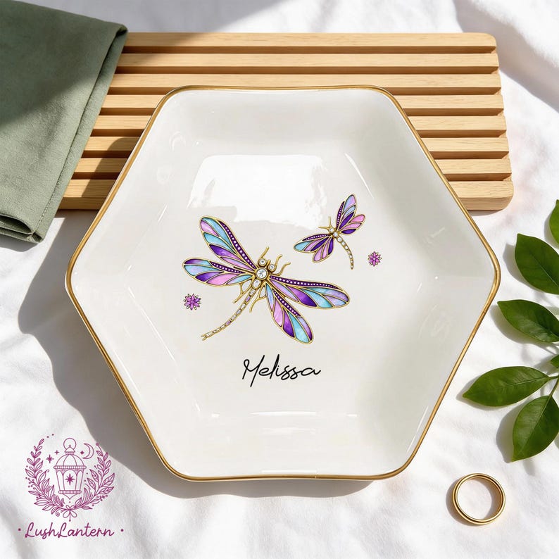 May include: White hexagonal trinket dish with a gold rim, featuring a dragonfly design in purple, blue, and gold. The name "Melissa" is written in script. A small gold ring is also visible.