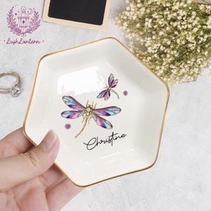 May include: White ceramic trinket dish with a gold rim, featuring a dragonfly design in purple, blue, and gold. The name "Christine" is written in cursive. The dish is held by a hand.