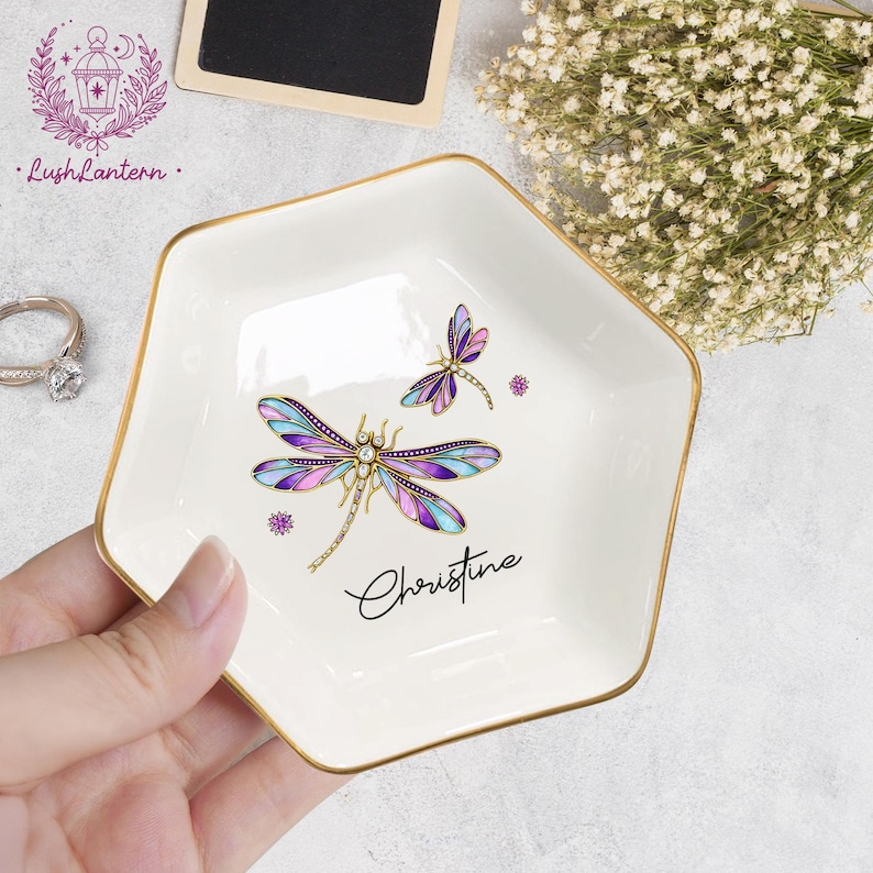 May include: White ceramic trinket dish with a gold rim, featuring a dragonfly design in purple, blue, and gold. The name "Christine" is written in cursive. The dish is held by a hand.