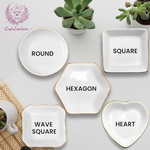 May include: A collection of white ceramic dishes in various shapes: round, square, hexagon, wave square, and heart. Each dish has a gold rim and a bold black label indicating its shape. The dishes are arranged on a light surface with small potted plants.
