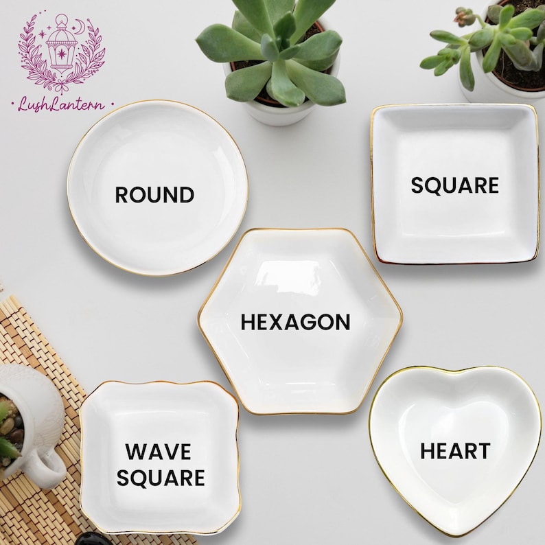 May include: A collection of white ceramic dishes in various shapes: round, square, hexagon, wave square, and heart. Each dish has a gold rim and a bold black label indicating its shape. The dishes are arranged on a light surface with small potted plants.