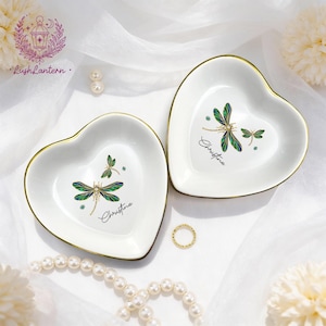 May include: Two heart-shaped white ceramic trinket dishes with gold rims. Each dish features a dragonfly design in green and blue, with the name "Christine" in script. A gold ring and pearl necklace are also visible.