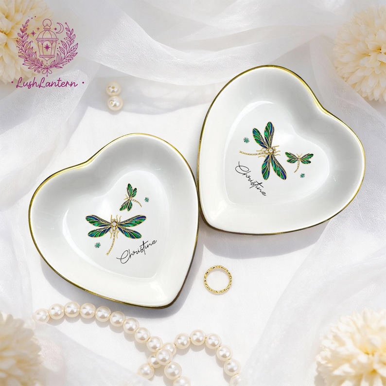 May include: Two heart-shaped white ceramic trinket dishes with gold rims. Each dish features a dragonfly design in green and blue, with the name "Christine" in script. A gold ring and pearl necklace are also visible.