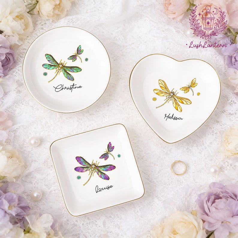 May include: Three white ceramic trinket dishes with gold rims. Each dish features a dragonfly design in shades of green, yellow, and purple, with a name printed below. The dishes are round, heart-shaped, and square.