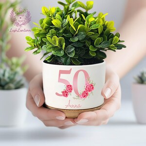 May include: A white ceramic planter with a green plant, featuring the number "50" in pink with floral accents and the name "Anna". The planter sits on a wooden base. The plant has vibrant green leaves.