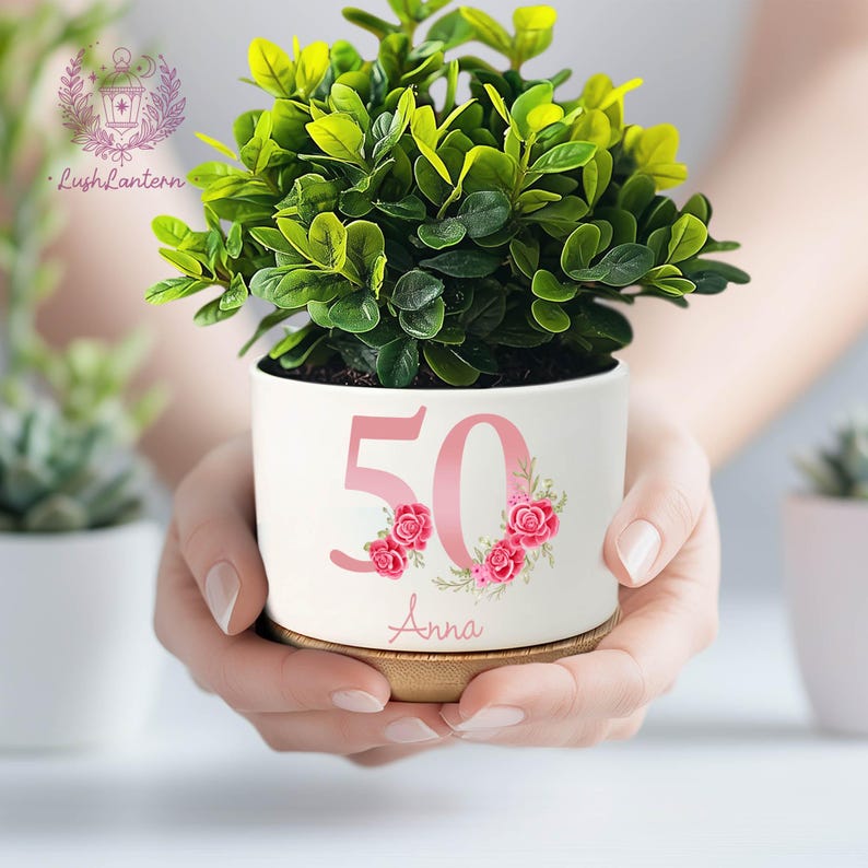 May include: A white ceramic planter with a green plant, featuring the number "50" in pink with floral accents and the name "Anna". The planter sits on a wooden base. The plant has vibrant green leaves.
