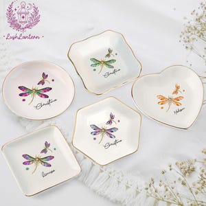 May include: A collection of small, decorative ceramic dishes in various shapes: round, square, heart, and hexagonal. Each dish features a dragonfly design with gold trim and a personalized name. The dragonfly wings are a mix of green, purple, and pink.