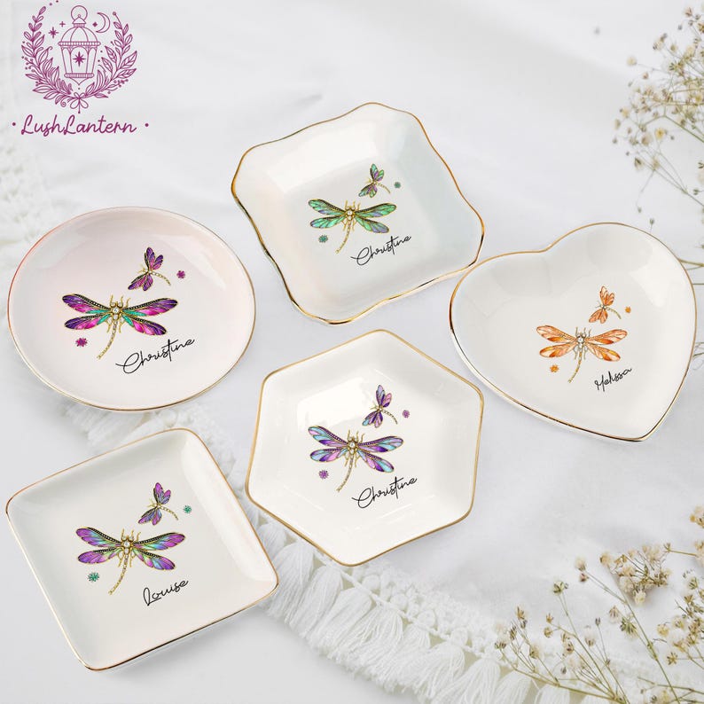 May include: A collection of small, decorative ceramic dishes in various shapes: round, square, heart, and hexagonal. Each dish features a dragonfly design with gold trim and a personalized name. The dragonfly wings are a mix of green, purple, and pink.