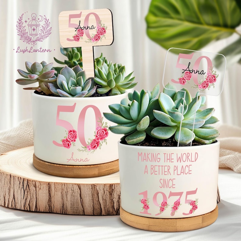 May include: Two white ceramic planters with succulents, each featuring a pink "50" with floral accents and the name "Anna." One planter has a wooden sign, the other a clear sign, both with matching design. The second planter reads "MAKING THE WORLD A BETTER PLACE SINCE 1975."
