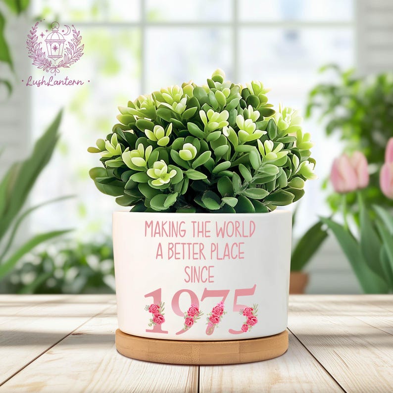 May include: A white ceramic planter with the text "MAKING THE WORLD A BETTER PLACE SINCE 1975" in pink. The planter is filled with a green plant and sits on a round wooden base. Floral accents decorate the numbers.