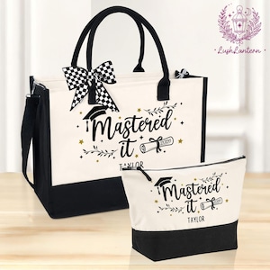 May include: A graduation-themed tote bag and matching cosmetic bag. The bags are off-white with black accents and handles. The text "Mastered It 2026" is printed on the front, along with a graduation cap and scroll. A checkered bow adorns the tote.