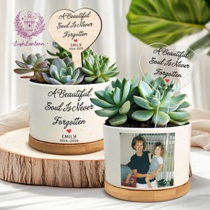 May include: Two white ceramic planters with succulents, each featuring a heart-shaped stake with the text "A Beautiful Soul Is Never Forgotten" and the name "EMILY 1954-2026". One planter has a photo. The planters sit on wooden bases.