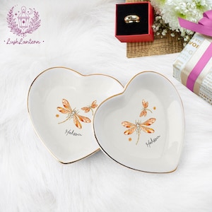 May include: Two heart-shaped, white ceramic trinket dishes with gold rims. Each dish features a dragonfly design in orange and brown, with the name "Melissa" written below. The dishes are on a white, fluffy surface.