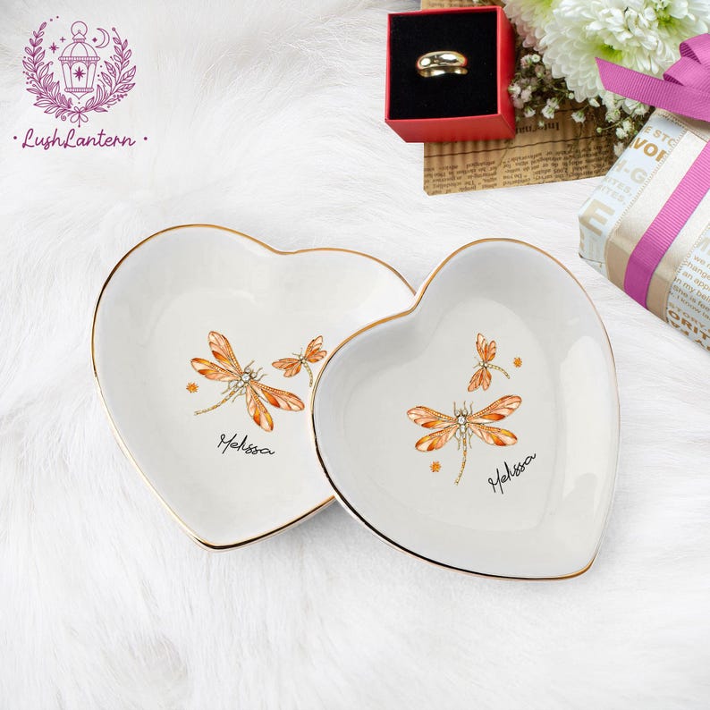 May include: Two heart-shaped, white ceramic trinket dishes with gold rims. Each dish features a dragonfly design in orange and brown, with the name "Melissa" written below. The dishes are on a white, fluffy surface.
