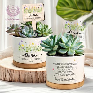 May include: Two white ceramic succulent planters with wooden bases. One planter has a wooden sign that reads "Olivia Officially Retired 2025". The other planter has a clear sign with the same text. Both planters have floral designs and succulents.
