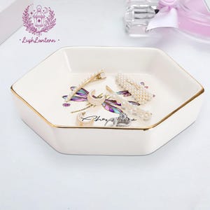 May include: A white hexagonal jewelry dish with gold trim, holding various accessories. The dish features a dragonfly design in purple, green, and gold. Accessories include pearl hair clips, a crescent moon hair clip, earrings, and a ring.