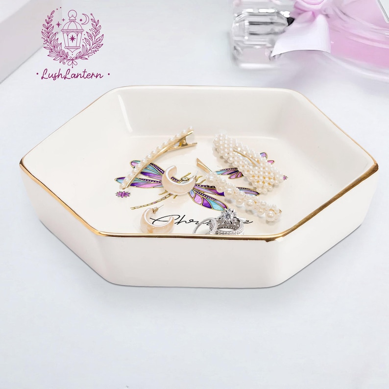 May include: A white hexagonal jewelry dish with gold trim, holding various accessories. The dish features a dragonfly design in purple, green, and gold. Accessories include pearl hair clips, a crescent moon hair clip, earrings, and a ring.