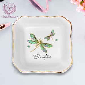 May include: White ceramic trinket dish with a gold rim and a dragonfly design. The dish features a large dragonfly with green, blue, and purple wings, and a smaller dragonfly. The name "Christine" is written in cursive.