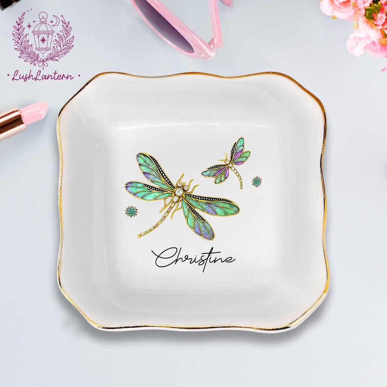 May include: White ceramic trinket dish with a gold rim and a dragonfly design. The dish features a large dragonfly with green, blue, and purple wings, and a smaller dragonfly. The name "Christine" is written in cursive.