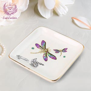 May include: A square, off-white ceramic trinket dish with gold trim. The dish features two dragonfly illustrations with purple, blue, and gold details. A diamond ring and a silver ring are on the dish. The word "muse" is written on the dish.
