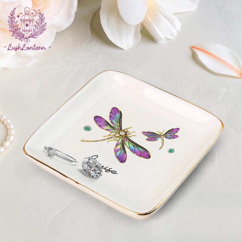 May include: A square, off-white ceramic trinket dish with gold trim. The dish features two dragonfly illustrations with purple, blue, and gold details. A diamond ring and a silver ring are on the dish. The word "muse" is written on the dish.