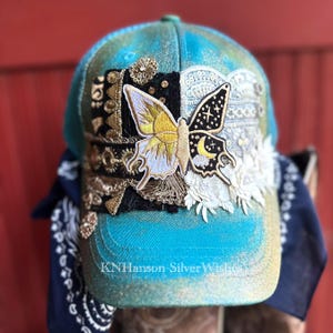 May include: A turquoise baseball cap with a gold-toned shimmer finish, featuring a central butterfly patch split between a yellow sun design and a black night sky with a moon. The cap is adorned with black lace, gold trim, and white floral lace.