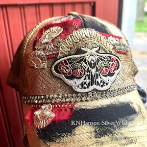 May include: Embellished trucker hat with a black and gold leopard print pattern. The hat features a large moth patch with red rose accents, gold embroidery, and rhinestone trim. The text "KNHanson-SilverWishes" is visible.