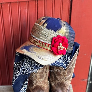May include: A custom baseball cap with a navy blue mesh back, gold and red embellishments, and a large red flower with a gold horse pin. The cap is paired with a navy blue bandana and brown cowboy boots.