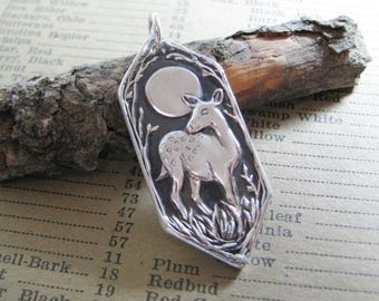 Puede incluir: Un colgante de plata con un diseño en relieve detallado de un ciervo de pie frente a una luna llena. El colgante tiene la forma de un hexágono con bordes redondeados.