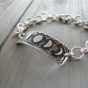 Puede incluir: Una pulsera de plata con un dije detallado de fases lunares. El dije presenta un dise&ntilde;o texturizado de &aacute;rboles y lunas crecientes.