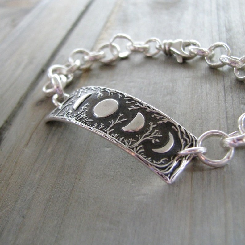 Puede incluir: Una pulsera de plata con un dije detallado de fases lunares. El dije presenta un dise&ntilde;o texturizado de &aacute;rboles y lunas crecientes.